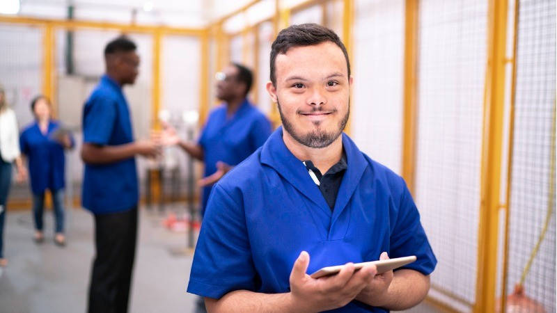 Un hombre con síndrome de Down y con un tablet sonríe en su trabajo, con otros compañeros al fondo