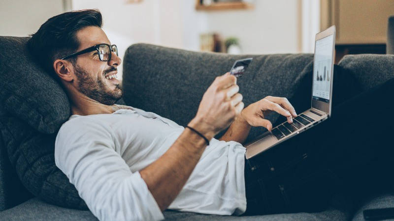 Medios de pago, autenticación reforzada; un hombre sonriente compra online con la tarjeta de crédito en la mano en el sofá 